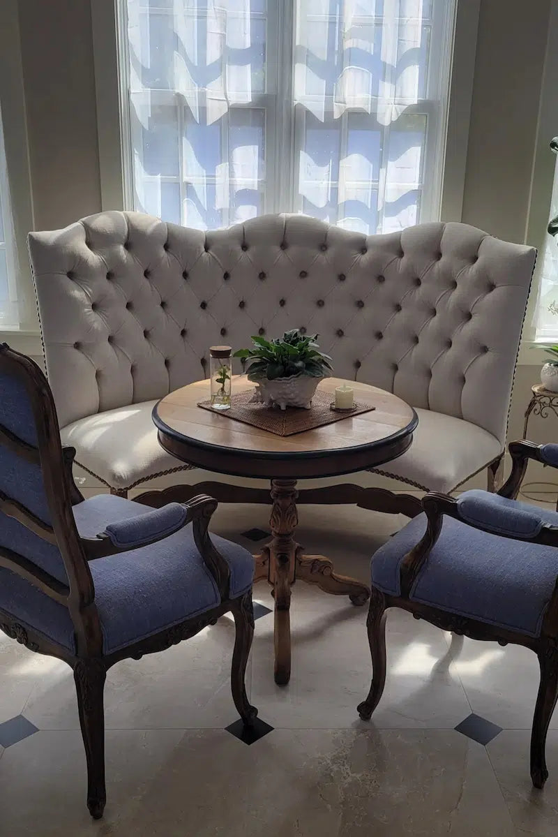Vintage-style seating area with a tufted white leather settee and blue lounge chairs in a room with large windows.